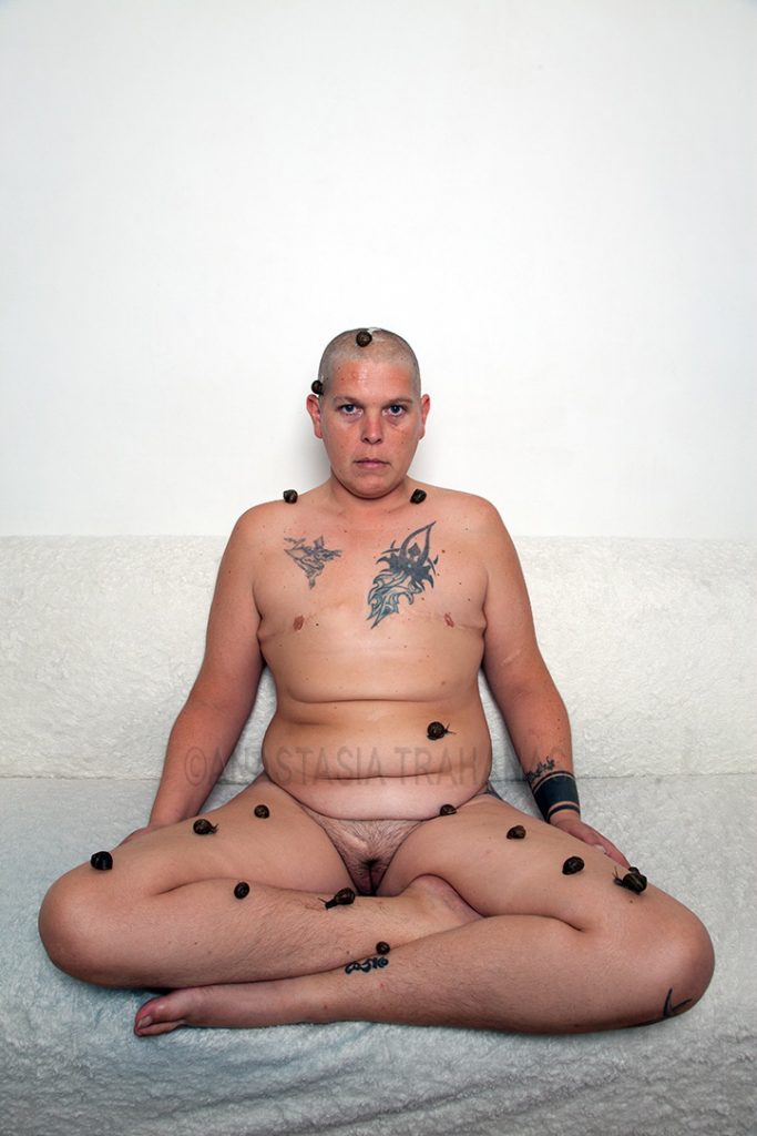 naked tattooed intersex with snails on him seated in yoga position on sofa