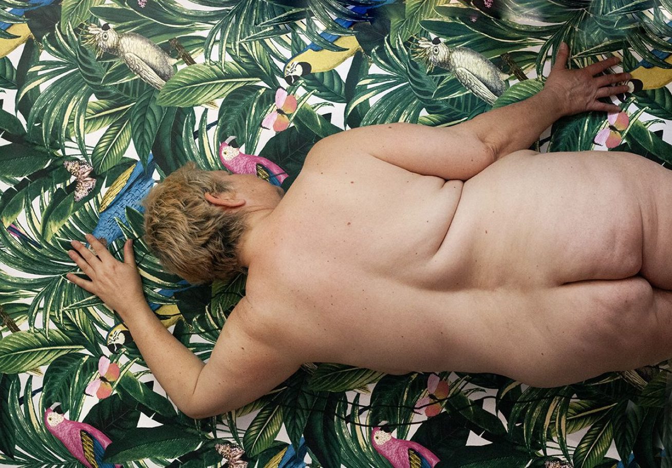nude photograph of a woman laying against forest print wall paper